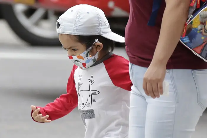 A child wearing a mask wearing a white cap holds the hand of an adult